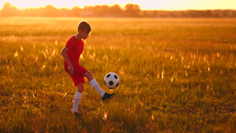 Niño-Junior-Con-Una-Camiseta-Roja-Y-Zapatillas-De-Deporte-Al-Atardecer-Haciendo-Malabarismos-Con-Un-Balón-De-Fútbol-Entrenando-Y-Preparándose-Para-Convertirse-En-Jugador-De-Fútbol.-El-Camino-Hacia-El-Sueño.-Trabajo-Duro